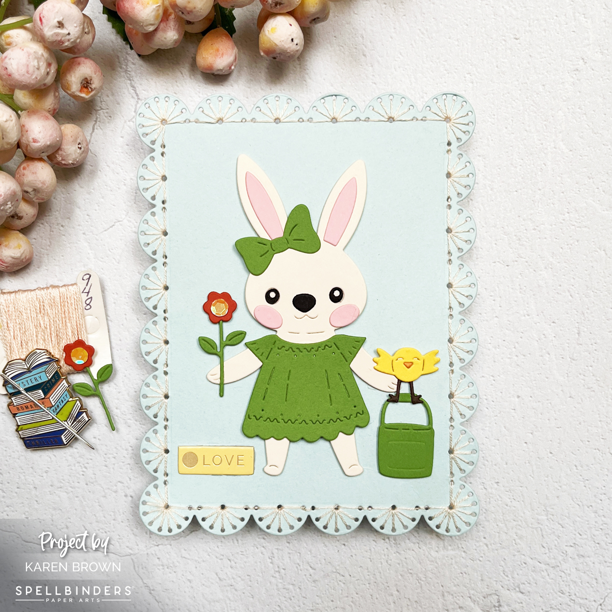 A handmade card featuring a die-cut bunny in a spring green dress holding a flower, framed with a stitched border. The background is soft blues and warm yellows, creating a fresh and cheerful scene.  Spellbinders Stitching Die of the Month + Large Bunny Die of the Month.