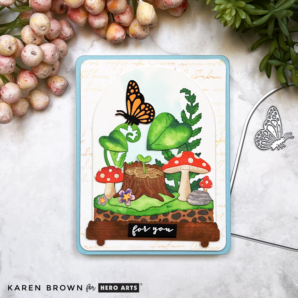 Botanical terrarium card  full of lush leaves, adorable red and tan mushrooms, green grass, rocks, a tree stump with a beautiful orange butterfly perched on a gently curling leaf.  I die cut my pieces from watercolor paper and then watercolored the components with brightly colored reactive inks.   Card created with Hero Studios April 2025 Card Kit of the Month