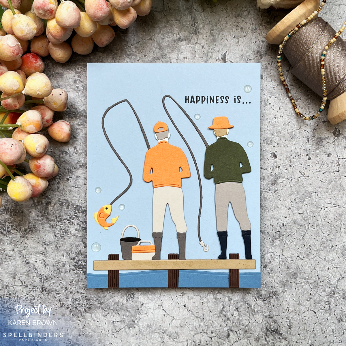 Handmade die-cut card featuring two male friends fishing on a wooden dock with fishing rods, tackle boxes, and a 'Happiness is...' sentiment. The color scheme is blue with pops of orange and green.