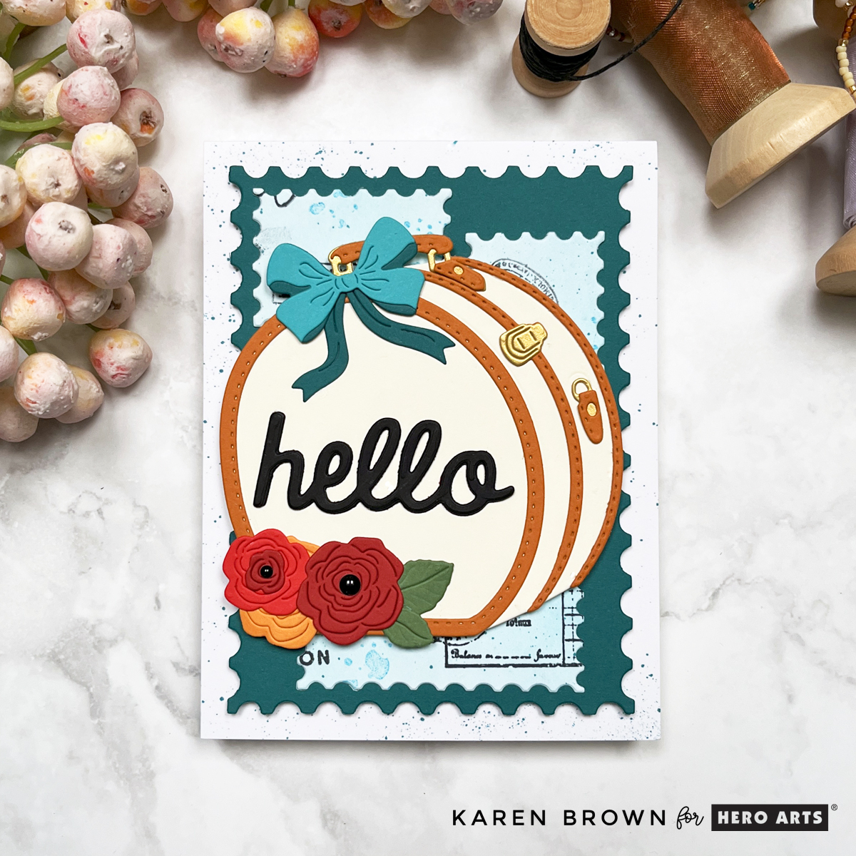 Hero Studio Suitcase and Florals die cut handmade "hello" card with a charming hat box luggage, teal bow, and flowers in orange and red. The background is a mixed media postage stamp theme.