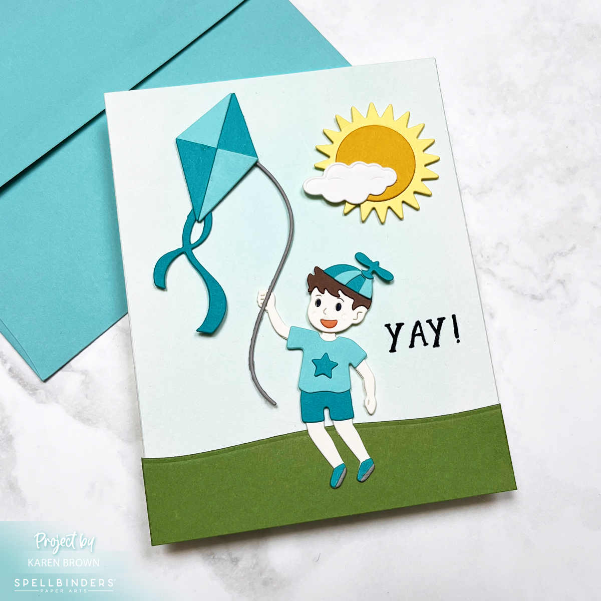 Handmade card featuring a die-cut boy flying a kite, with a die-cut sun and cloud above. Background is a soft aqua watercolor wash. Card uses teal and aqua cardstock in a playful, kite and sky-themed scene.