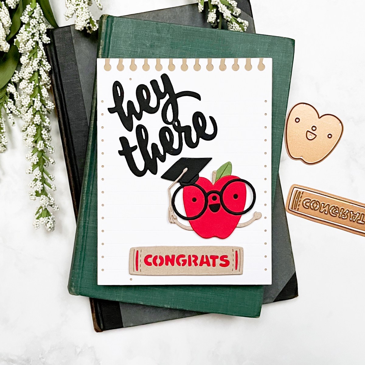Handmade card featuring a smiling die-cut apple with glasses and a graduation cap on a white memo paper background. Includes a “hey there” sentiment and a “congrats” die shaped like a book spine. Color scheme: red, kraft, and black.