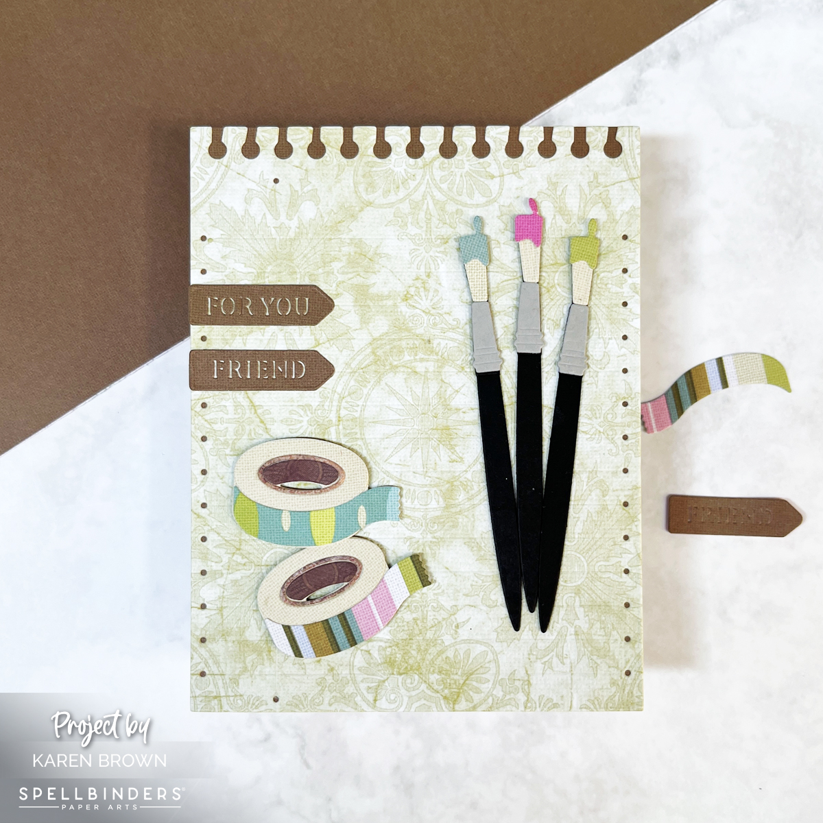 Craft theme greeting card with three  die-cut paint brushes, two rolls of die cut patterned Washi tape and a die cut Notebook background in muted greens and browns.