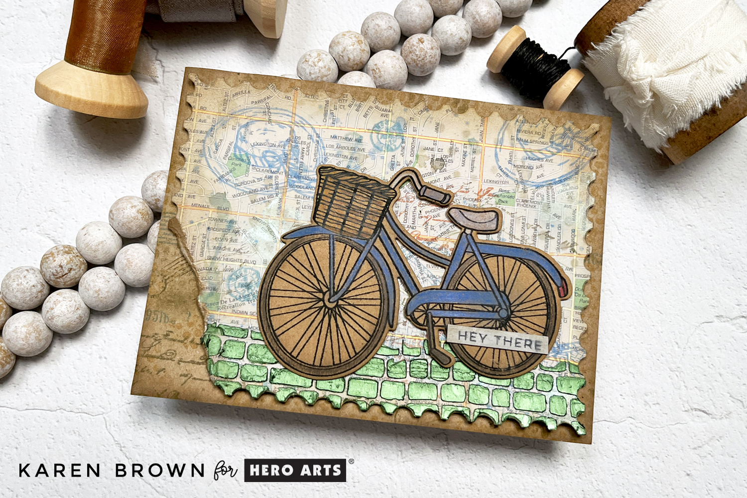 How to tutorial for a tattered and torn mixed media travel theme card with a die cut bike, upcycled road atlas background and stenciled Hero Wax accent.