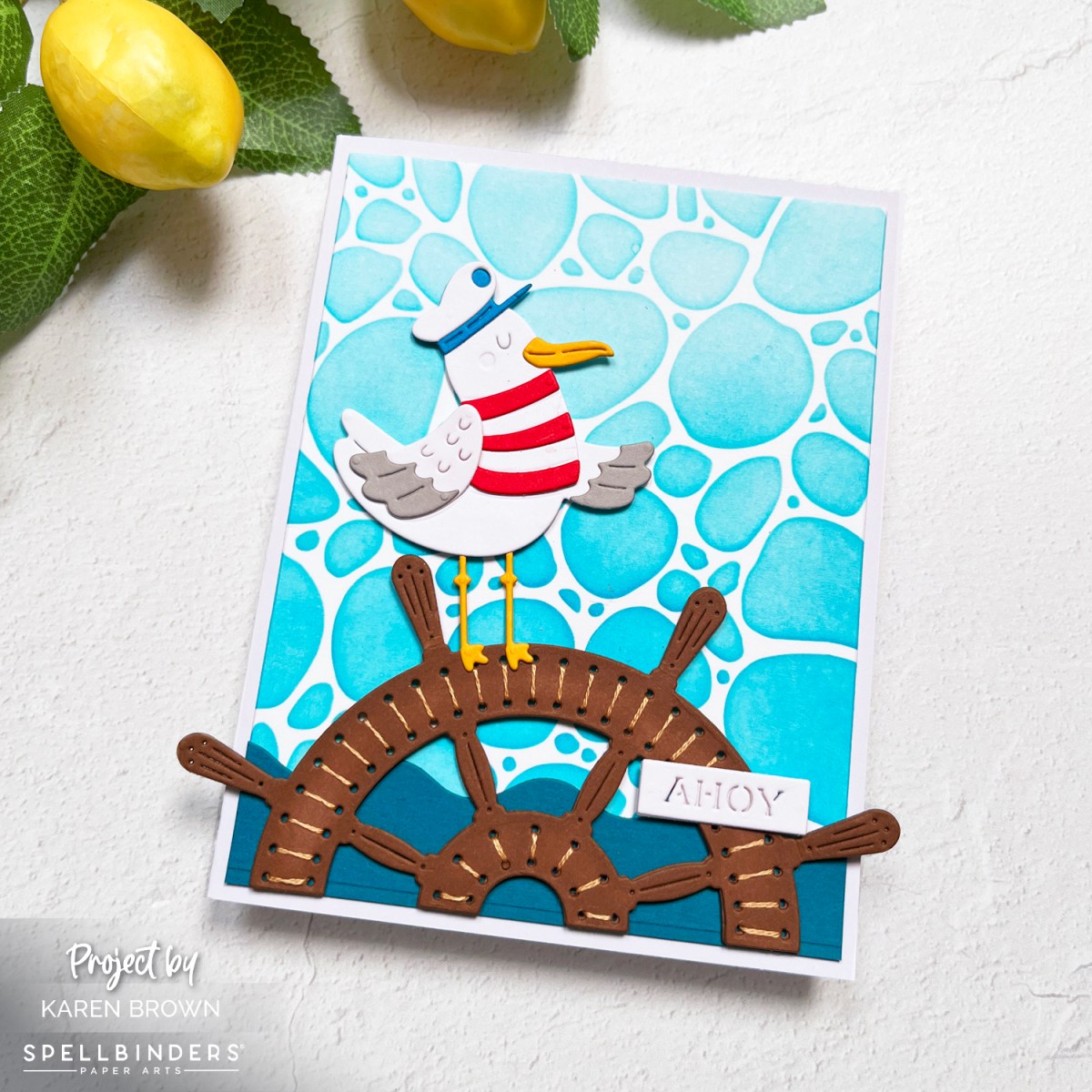 Card with a Stitched captain's wheel on an aqua stenciled background with a seagull in a red and white striped shirt and “ahoy” sentiment.  Spellbinders June 2025 Stitching Die of the Month Kit.