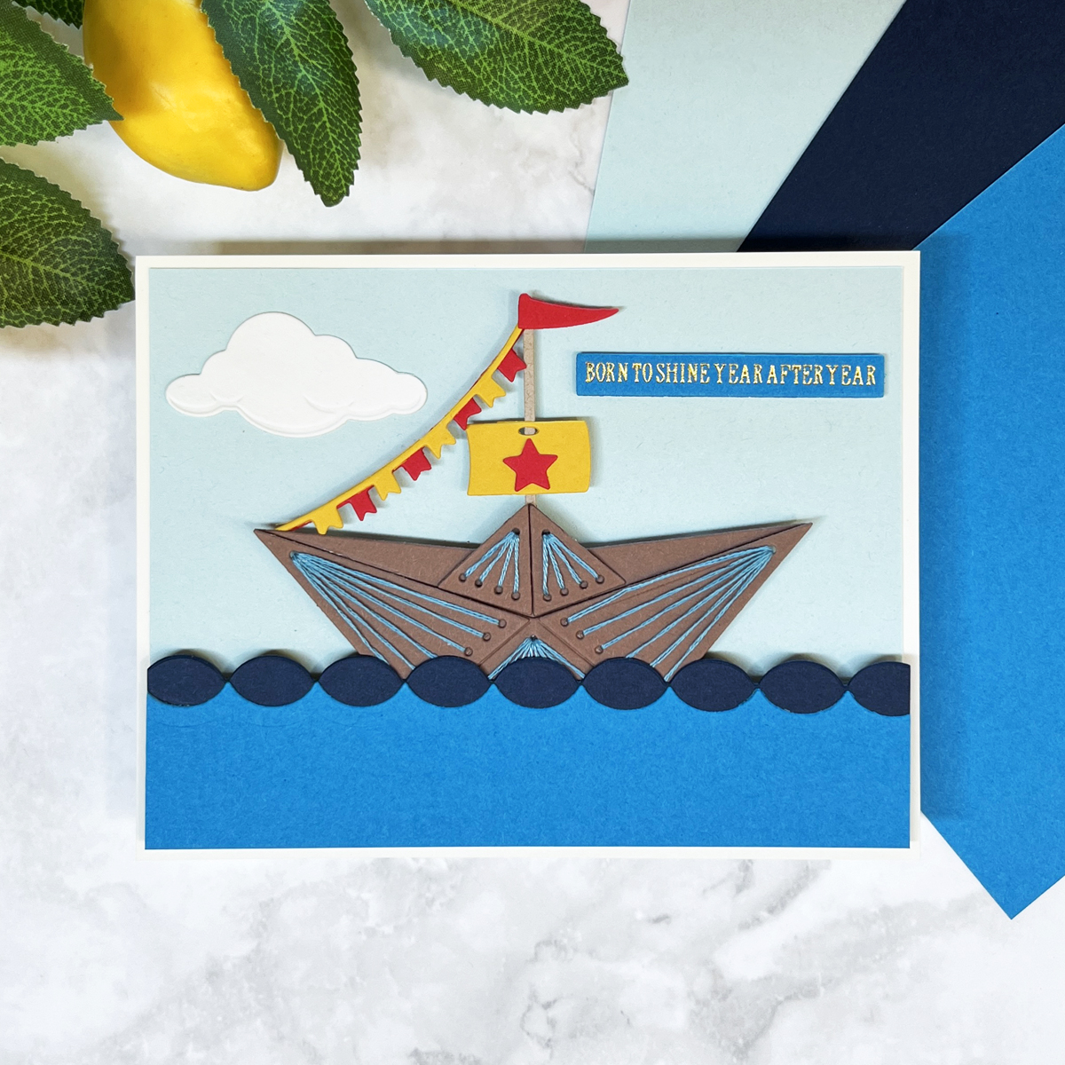 Stitched and die cut brown sailboat with red and yellow flags on blue sea and “Born to Shine Year After Year” sentiment. Made with Spellbinder's August 2025 Stitching Die of the Month.