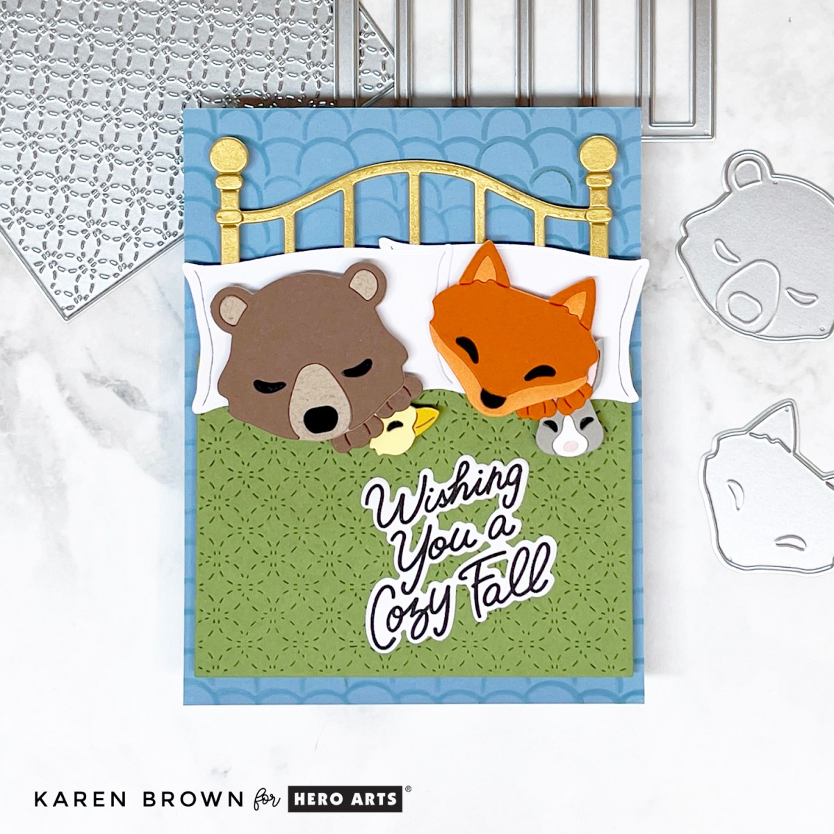 Die cut scene card featuring a brown bear and orange fox tucked into a brass bed with a green quilt. Bear snuggles a duck, fox holds a bunny. Sentiment “Wishing You a Cozy Fall” with a subtle stenciled wallpaper background.
