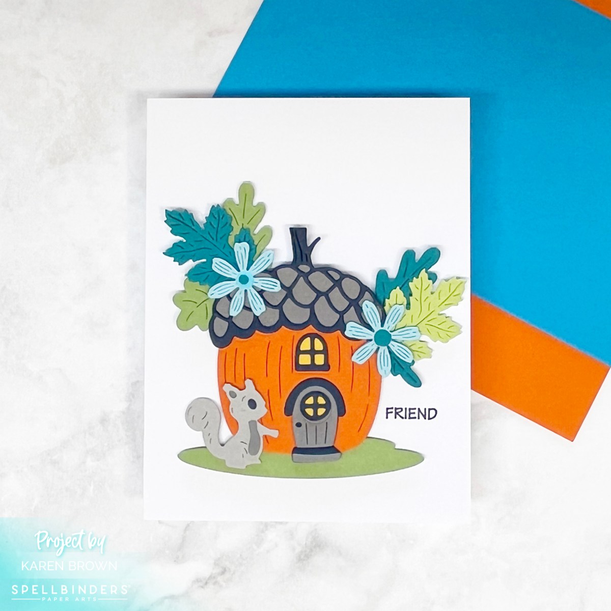 Die cut "pumpkin" house card, with a gray squirrel walking toward the door. The roof is decorated with teal and green leaves and two aqua flowers. 