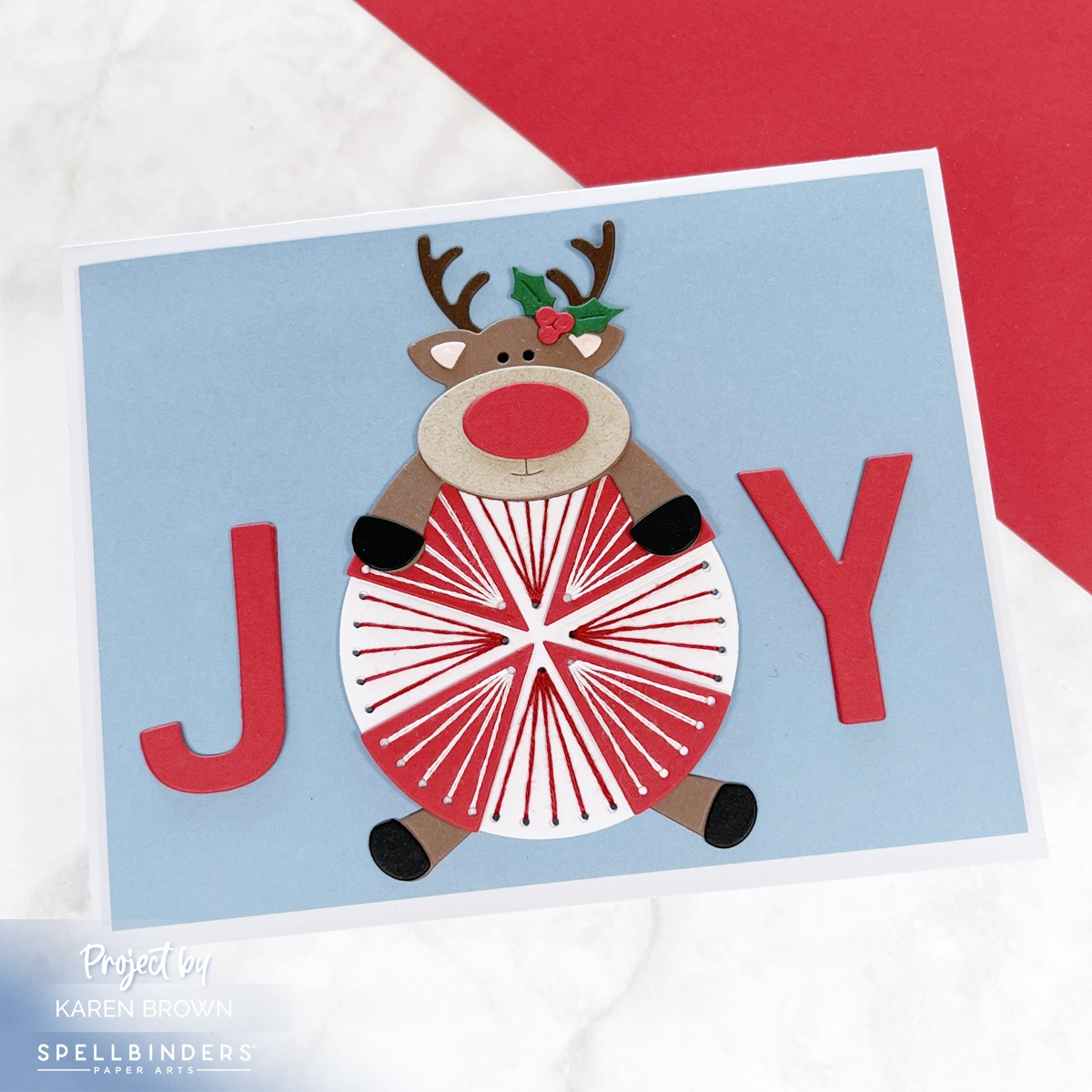 Spellbinders October 2025 Stitching Die of the Month handmade Christmas card with a stitched red and white peppermint candy used as the O in JOY, with a playful Rudolph reindeer sprawled across it on a Cascade blue background
