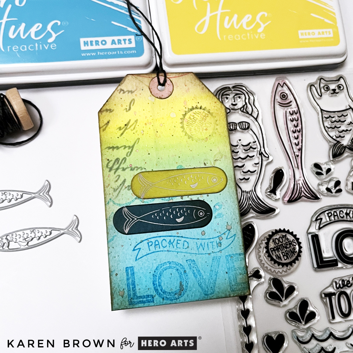 Close-up of mixed media tag featuring layered stamping, script texture, inked edges, and a foiled fish accent on a colorful watercolor background.