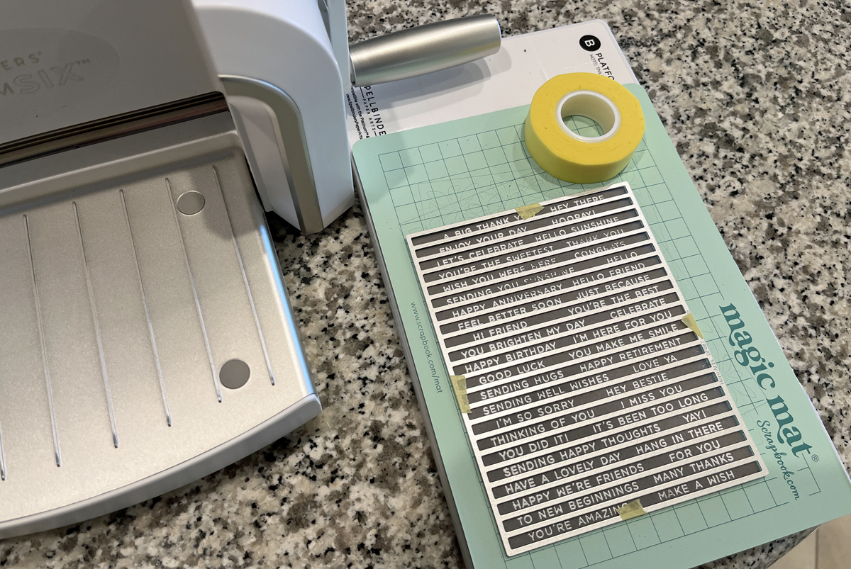 Process photo of heat embossed sentiment strip panel in white embossing powder on gray cardstock with coordinating sentiment strip die taped in place, ready for die cutting.