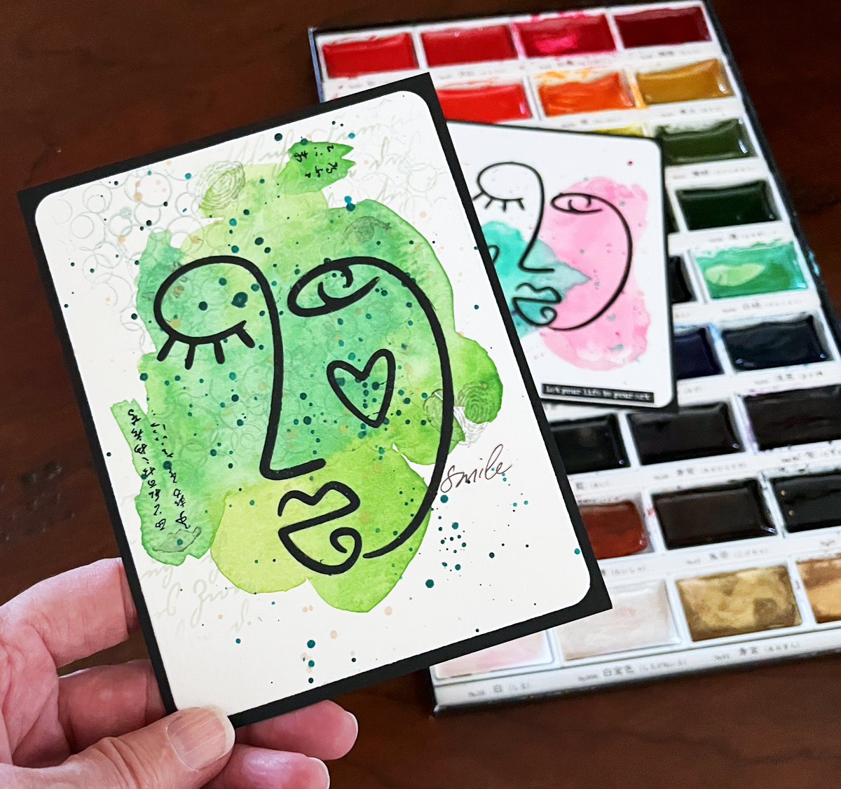 Mixed media card with green watercolor face background and black abstract face die cut with a second version in the background with my watercolor paint palette.