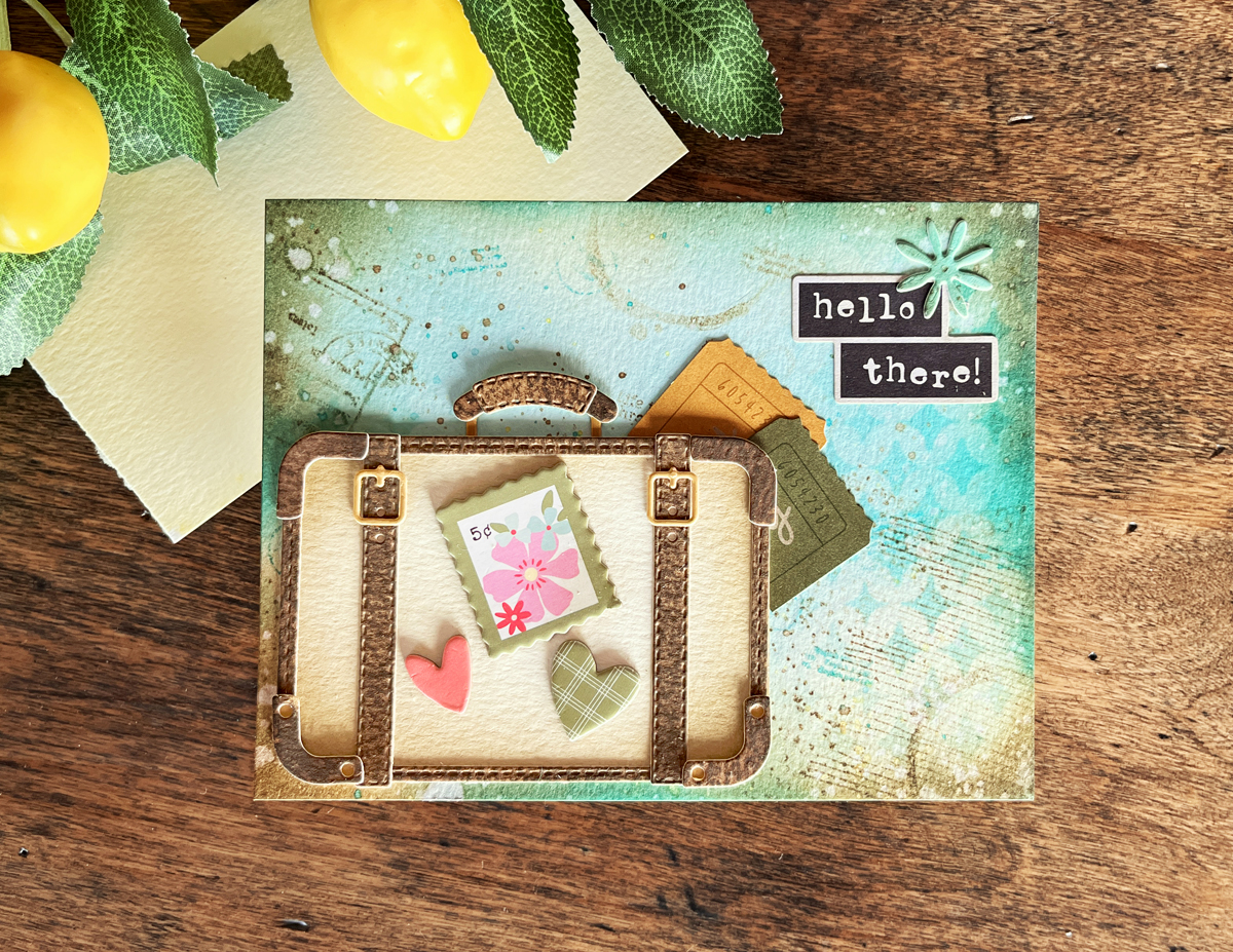 Mixed media card featuring layered ink blended background with stenciling, stamping, splatters, and a vintage suitcase focal point in aqua, yellow, and brown tones.