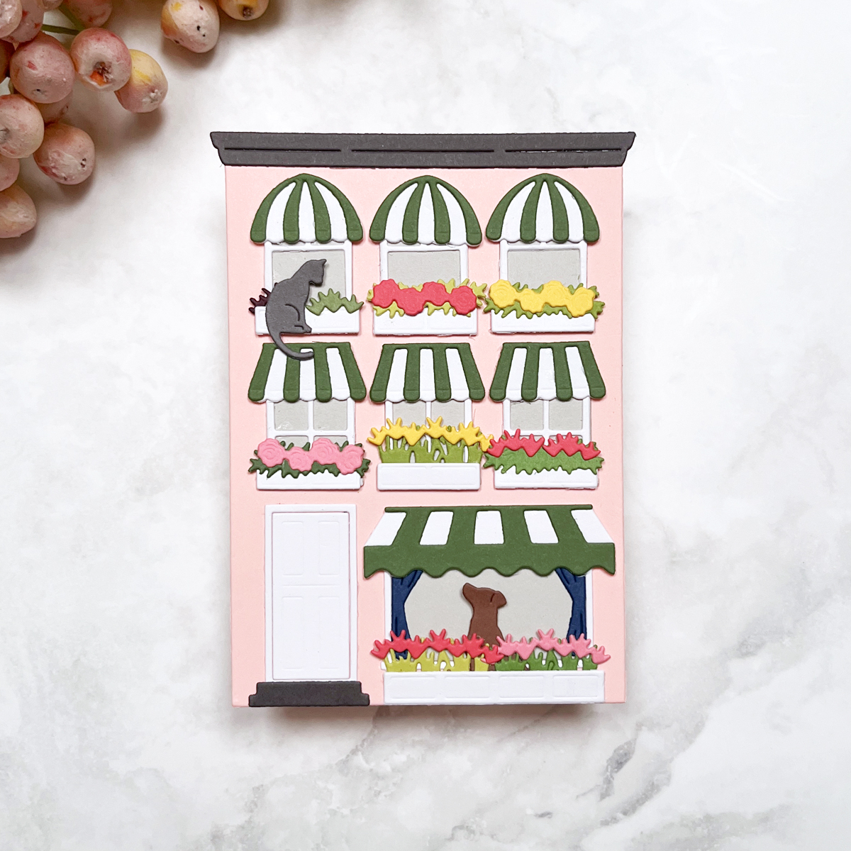 Die cut house scene card with pink building, green striped awnings, white windows, and colorful flower boxes