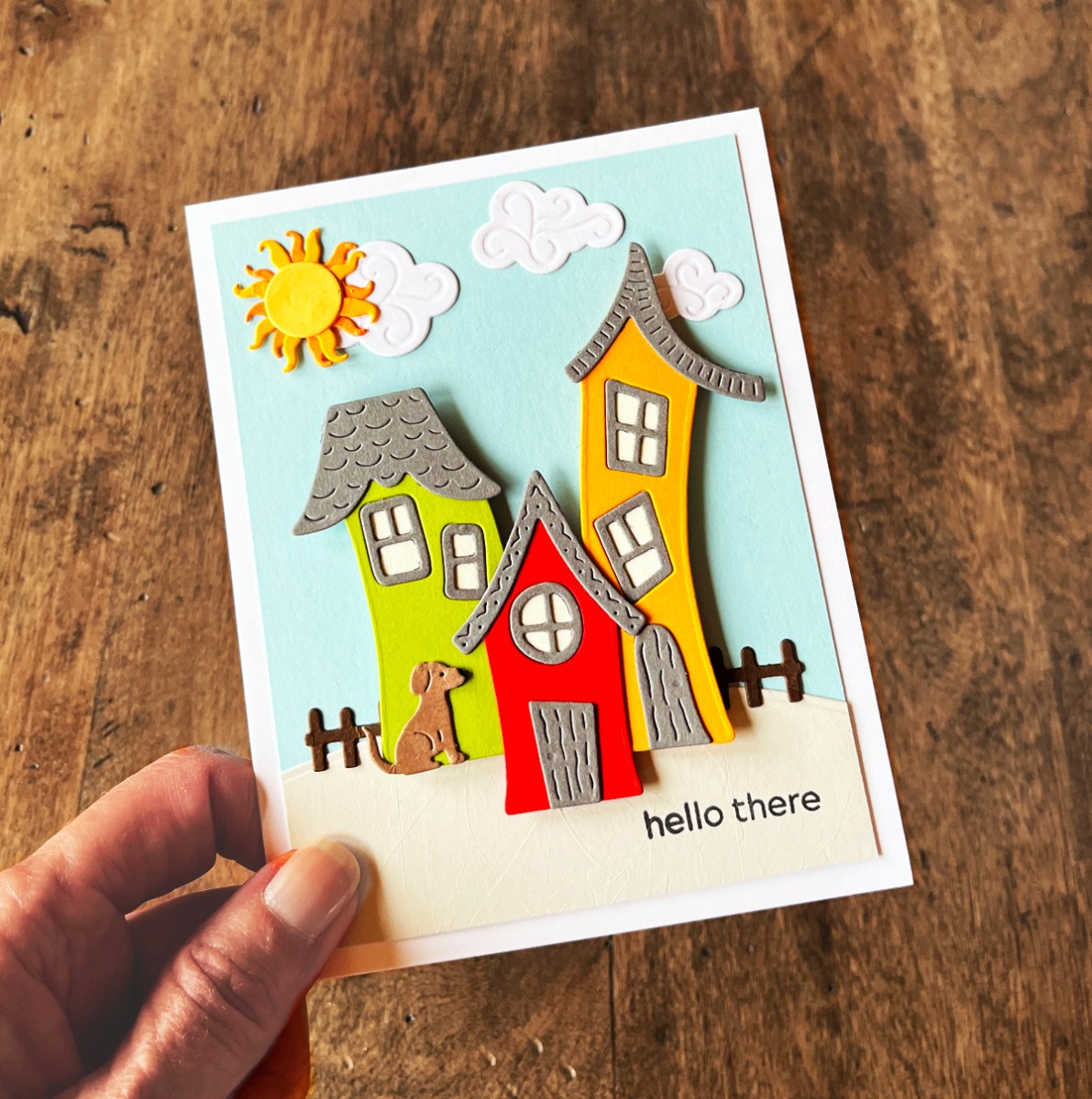 Whimsical die cut village scene card featuring colorful storybook-style houses, clouds, sun, and dimensional layered details on a blue background.