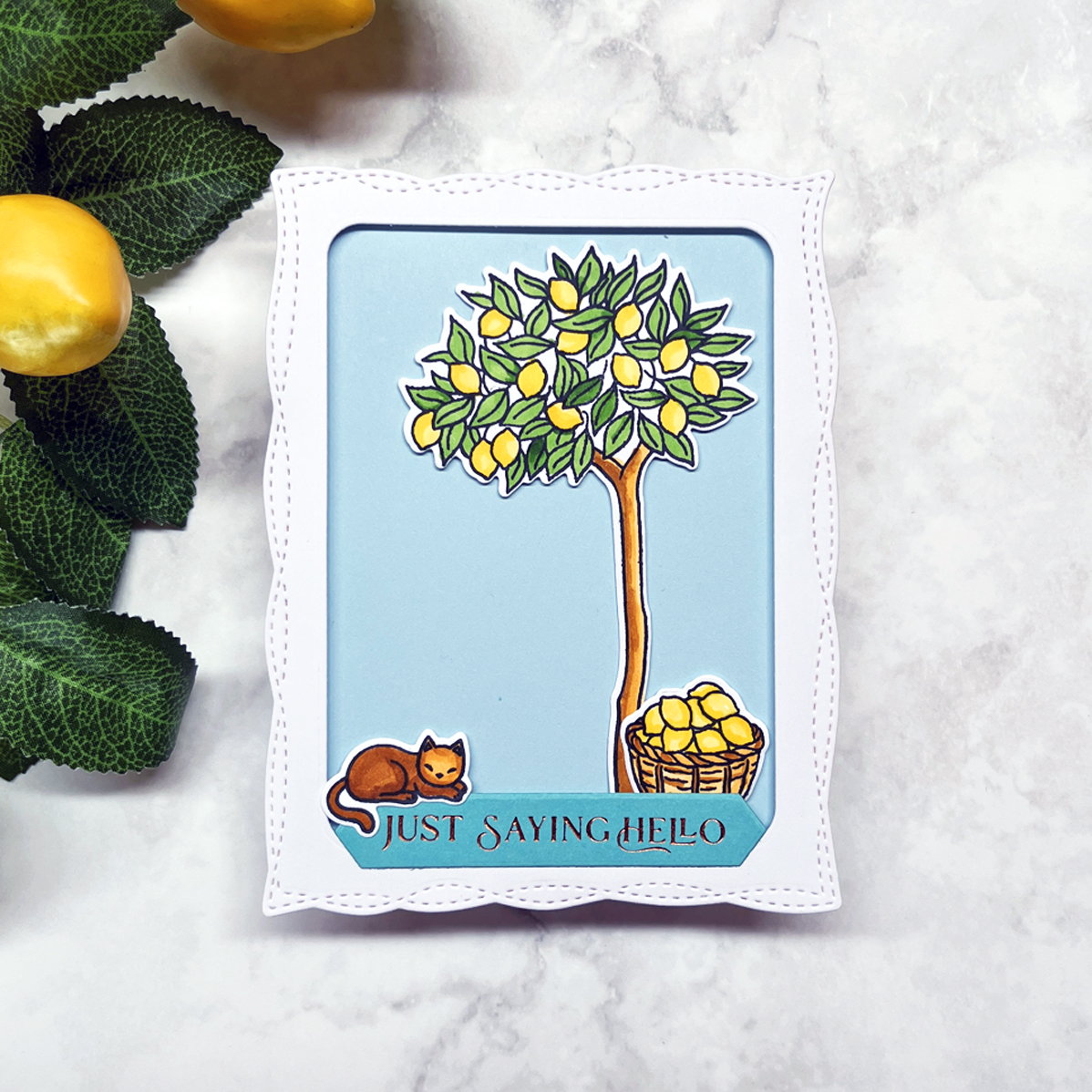 Stamped and colored lemon tree scene card featuring a lounging cat, basket of lemons, stitched window frame, and soft blue background.