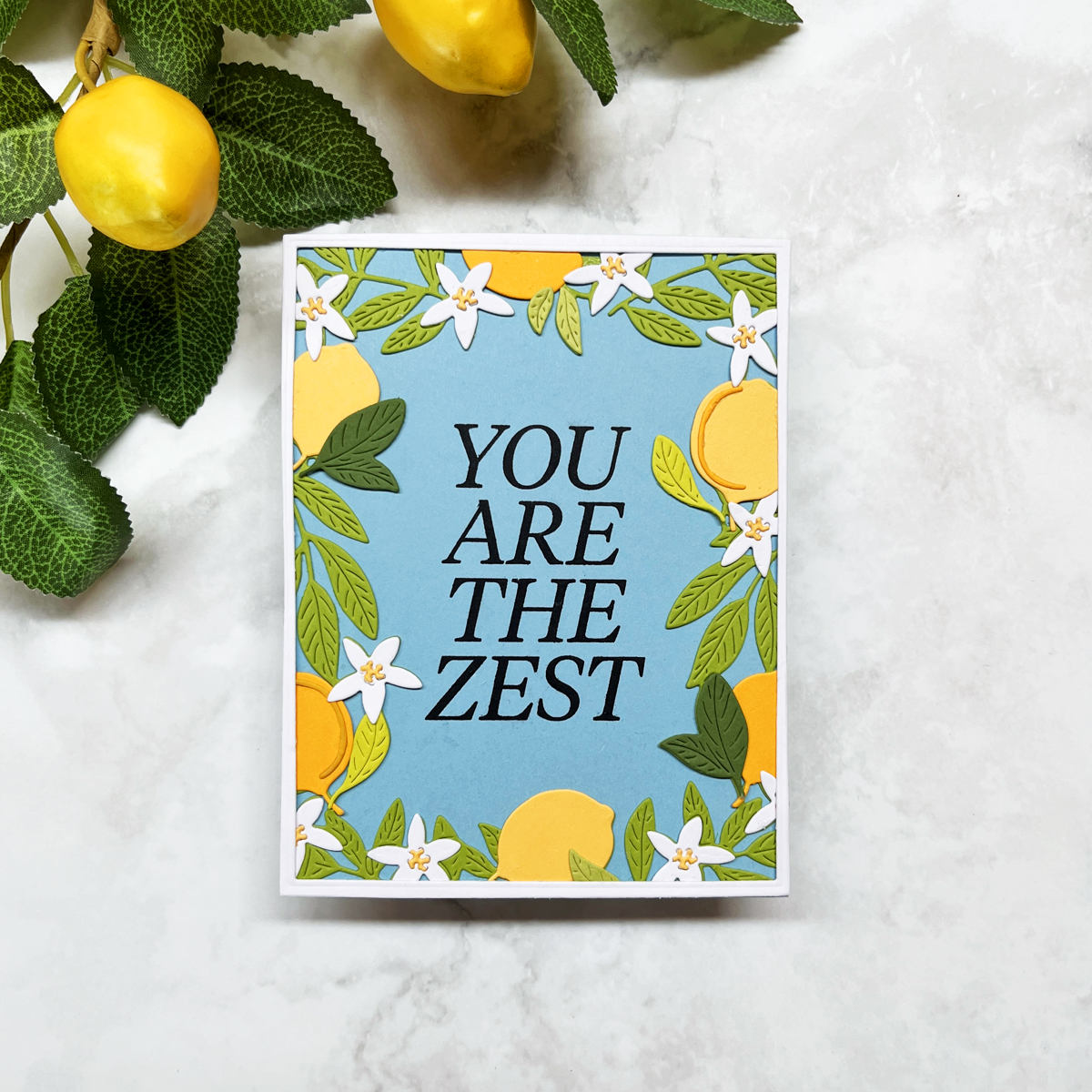 Layered die-cut citrus frame card with yellow lemons, white blossoms, green leaves, and a large "You Are the Zest" sentiment on a soft blue background.