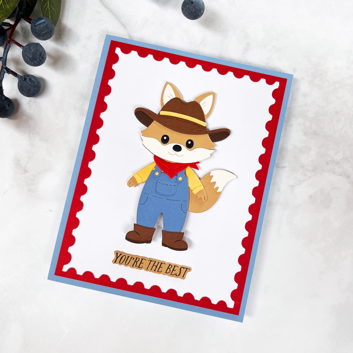 Handmade animal card featuring a tan die-cut fox dressed as a rancher in blue overalls, cowboy hat and red kerchief, framed with white, red and blue cardstock.