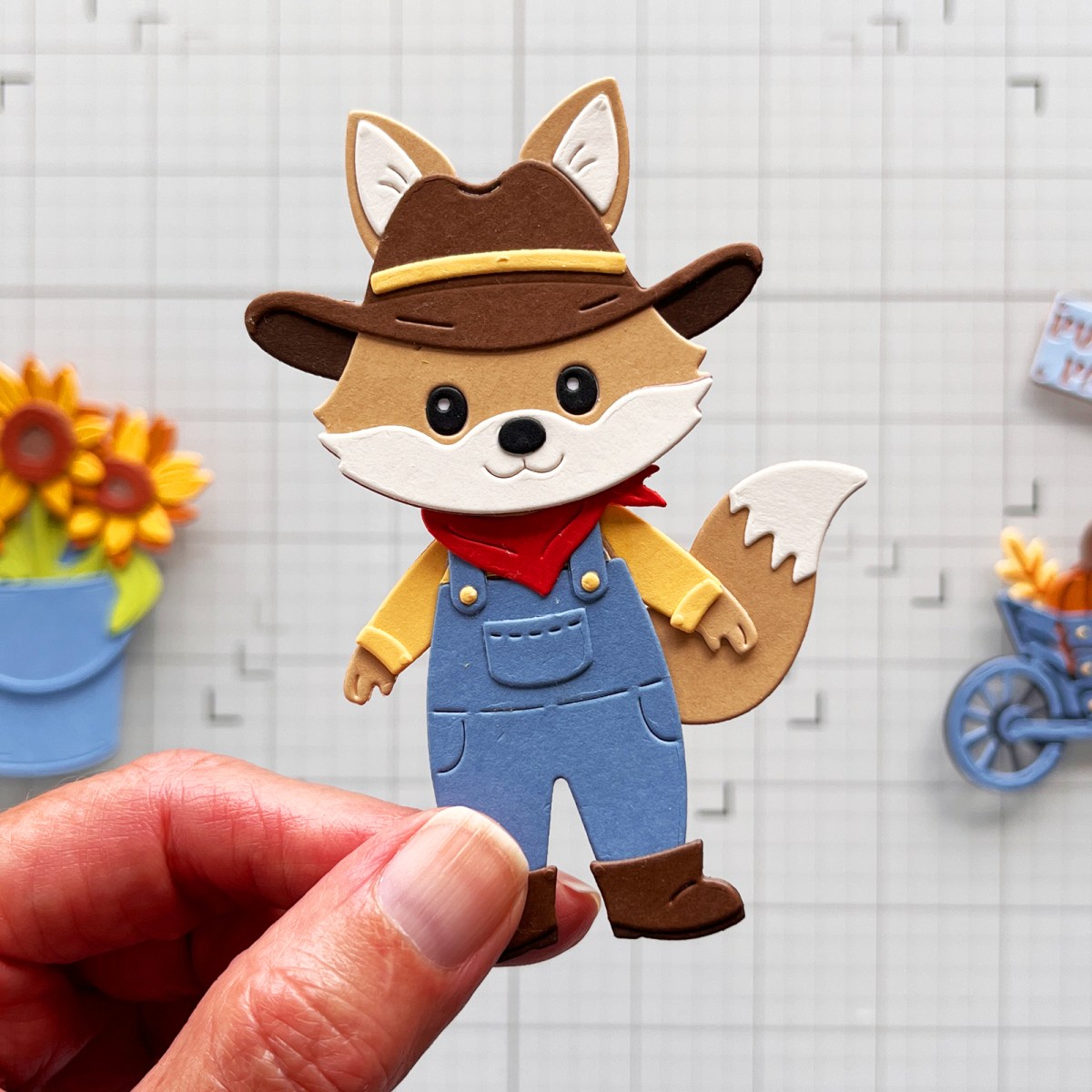 Hand holding an assembled tan die-cut fox dressed in blue overalls, yellow shirt, red kerchief and cowboy hat over a craft mat.