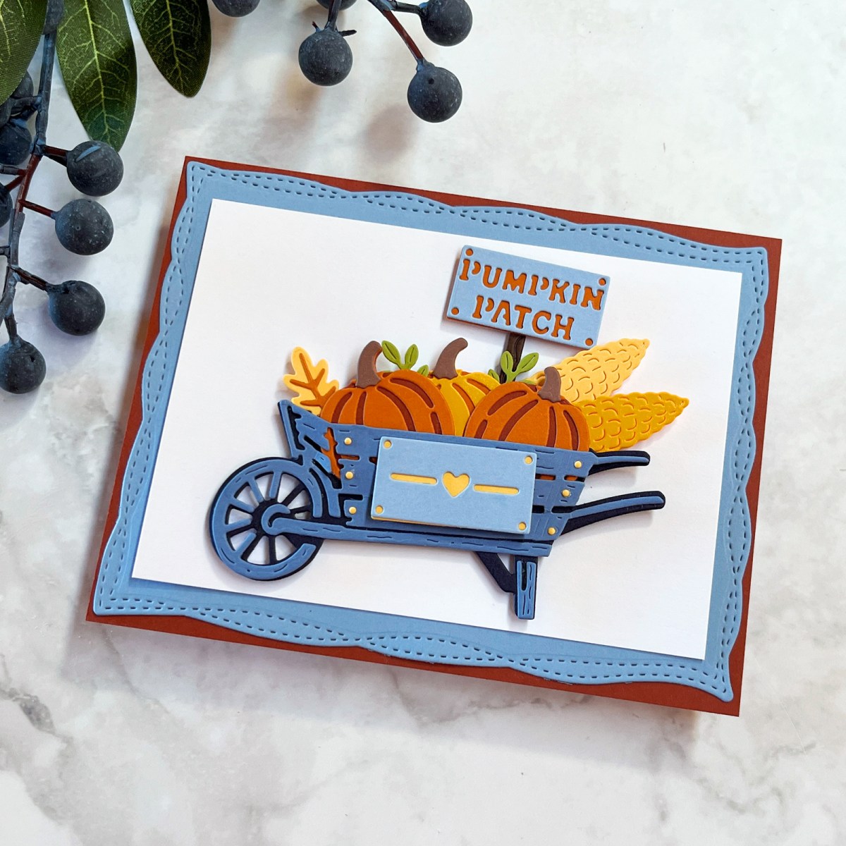 Rustic wheelbarrow card featuring die-cut pumpkins and corn in a blue, orange and yellow color scheme with layered stitched background.  