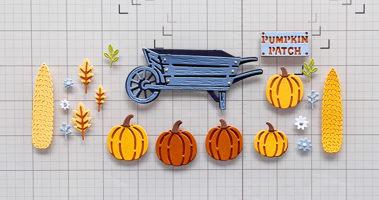 Blue die-cut wheelbarrow, pumpkins, corn, flowers and leaves arranged on a craft mat before assembling the card focal point.
