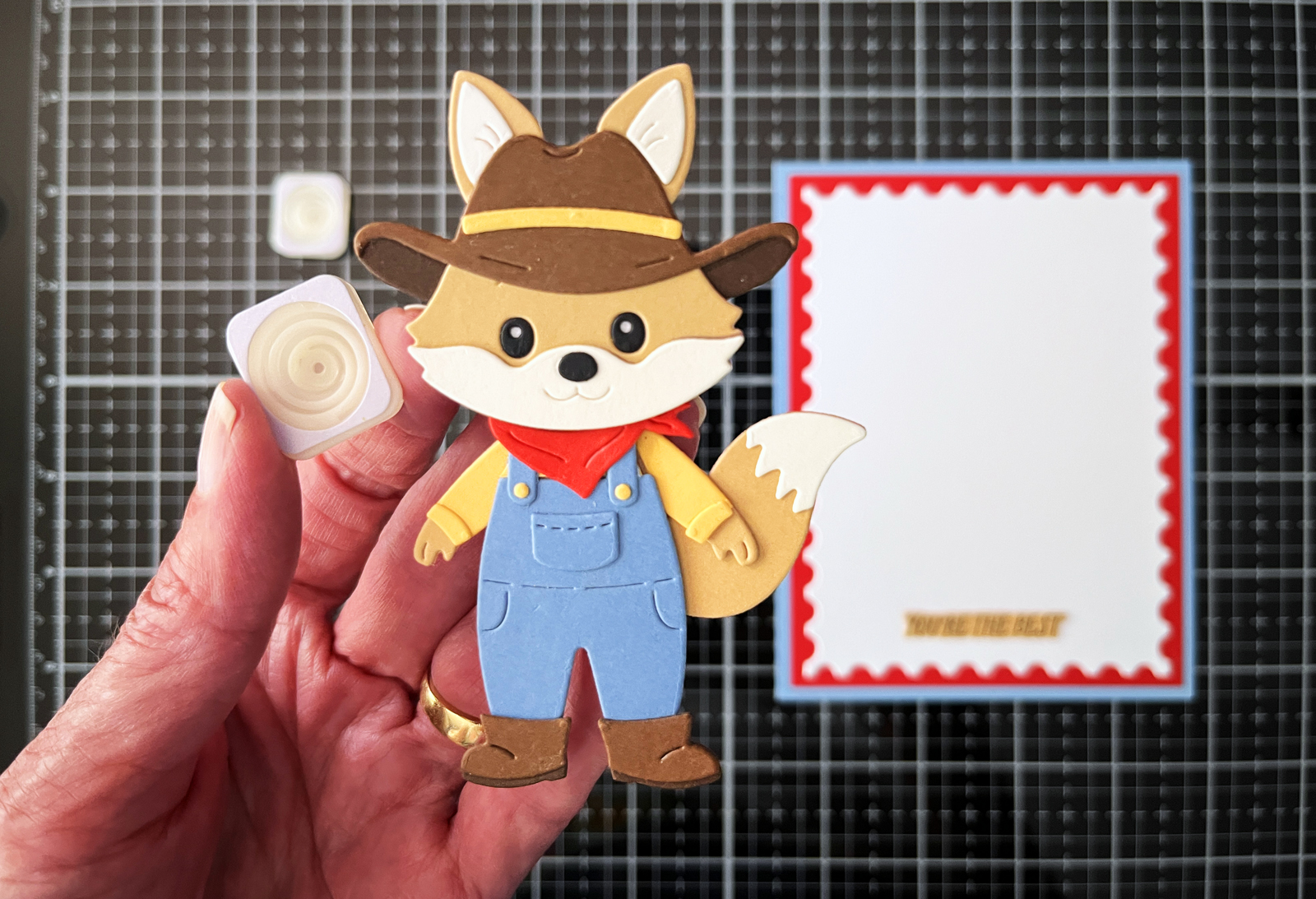 Hand holding a die-cut rancher fox and Giggle Spring over a craft mat with the layered card background and another spring in view